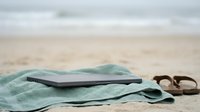Young adult male of South Asian ethnicity relaxing on sandy beach with tablet device and sunglasses on turquoise towel during summer vacation.