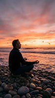 Man meditating in lotus position on pebble beach at sunset with dramatic orange pink sky and seagulls flying overhead creating peaceful mindfulness scene.