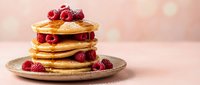 Stack of golden pancakes topped with fresh raspberries and syrup on plate against soft pink bokeh background for breakfast menu design.