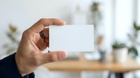 Person with medium skin tone holding blank white business card in modern office environment with wooden desk and plants.