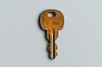 Old brass house key with worn patina and distinctive ridged pattern photographed on light gray background, isolated view showing security details.