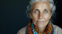 Senior woman with striking blue eyes wearing colorful ethnic scarf and beige sweater against dark background, natural aging beauty portrait showing wisdom and grace.