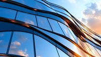 Modern curved glass building facade with dramatic sunset reflection creating dynamic pattern of light and shadow against blue sky with clouds.