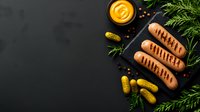 Grilled sausages served on black slate board with mustard sauce, pickled cucumbers, fresh herbs and spices on dark background. Perfect for menu design.