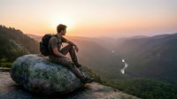 Male hiker with backpack sitting on rocky cliff overlooking misty mountain valley at sunrise, contemplating scenic landscape view for travel and adventure concepts.