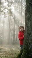 Young child in red jacket peeking around large tree trunk in misty forest with bokeh sunlight filtering through trees, suitable for children's books, parenting content, and nature editorial use.