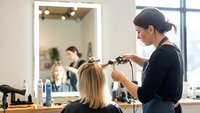 Professional female hairstylist with dark hair in bun styling blonde client's hair with curling iron in modern bright salon with mirror and window.