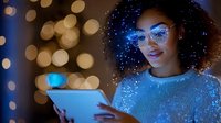 Young African American woman with curly hair and glasses illuminated by blue light from digital tablet against bokeh background with magical sparkle effects.