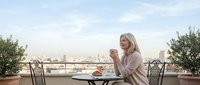 Young blonde woman enjoying morning coffee on urban rooftop terrace with city skyline view, relaxing breakfast moment on balcony with croissants and peaceful atmosphere.