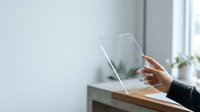 Female hand with medium skin tone holding transparent glass tablet device on white desk with blurred office background, suitable for technology, business, and digital innovation concepts.