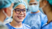 Young female medical professional in scrub cap and glasses smiling during surgical procedure, showing teamwork and positive attitude in healthcare setting.
