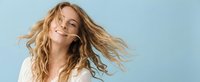 Young blonde woman with flowing wavy hair in motion against soft blue background, expressing joy and freedom with natural smile and carefree movement.