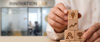 Businessman stacking wooden blocks with innovation icons including lightbulb, arrow, gears and trophy in modern office environment for business growth concepts.