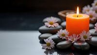 Orange candle burning surrounded by smooth black spa stones and white daisy flowers creating peaceful zen meditation atmosphere for wellness relaxation.