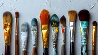 Colorful used artist paintbrushes in a row showing texture and wear from creative use, with paint stains and golden details against light textured background.