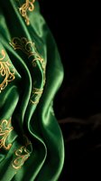 Luxurious emerald green silk fabric with golden ornate embroidery flowing in elegant folds against dark background for premium textile and fashion design.