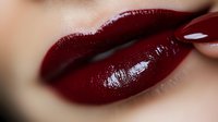 Close up of sensual dark burgundy glossy lips with matching manicured nail touching them, showing luxury cosmetics and beauty makeup artistry in soft lighting.