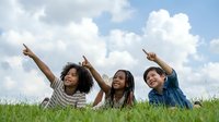 Three diverse young women lying on grass pointing upward with joyful expressions against cloudy blue sky background for lifestyle and friendship concepts.