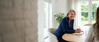 Mature woman with blonde hair, Caucasian ethnicity, wearing a navy blazer, seated at a wooden table in a bright office space with large windows overlooking greenery, engaged in professional discussion or interview.