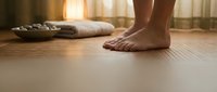 Barefoot person standing on wooden floor in warm sunlit room with folded towels and curtains, wellness and home relaxation concept.