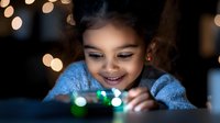 Young South Asian girl smiling while playing with glowing lights, wearing blue lace dress against dark background with soft bokeh effect, creating magical atmosphere.