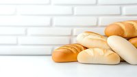 Fresh baked white and wheat submarine sandwich rolls stacked on white wooden table against light brick wall background, food photography.