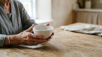 Senior woman with weathered hands holding warm ceramic bowl at wooden table, wearing gray cardigan in bright home interior with soft natural light, conveying comfort and wellness.
