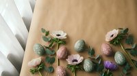 Colorful speckled Easter eggs with pink anemone flowers and eucalyptus branches arranged on kraft paper background for spring holiday celebration decor.