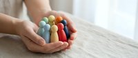 Hands gently holding colorful wooden peg people figures representing diversity, inclusion, teamwork and community support in workplace or social setting.