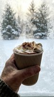 Steaming hot chocolate with toasted marshmallows and whipped cream held by hand against snowy winter forest window backdrop.