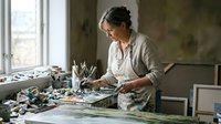 Female artist painting in bright studio workspace with natural light from large windows surrounded by art supplies and creative materials.