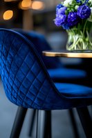 Luxurious royal blue quilted velvet bar stool with black legs and purple flowers in glass vase on golden table, elegant interior design detail with bokeh background.