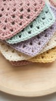 Colorful crocheted granny squares in pastel pink, mint green, lavender and cream yarn stacked on wooden surface for handmade craft projects and textile art.
