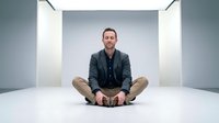 Professional Caucasian businessman in dark blazer sitting cross-legged in modern minimalist white interior space for corporate wellness and mindfulness concepts.