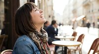 Happy young woman laughing outdoors at street cafe wearing denim jacket and patterned scarf. Joyful moment of urban lifestyle and positive emotions.