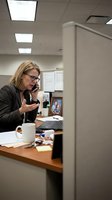 Professional businesswoman talking on phone at office cubicle desk with laptop and family photo, corporate workplace environment for business communications.
