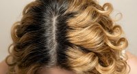Blonde highlighted curly hair with dark roots showing natural regrowth and center part styling on woman's head against neutral background.