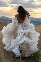 Young woman in flowing white ruffled dress stands against mountain sunset backdrop, her dark hair blowing in wind as dramatic fabric billows around her in golden field.