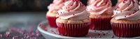 Red velvet cupcakes with pink cream cheese frosting swirls decorated with heart sprinkles on white plate against dark background, romantic dessert setup.