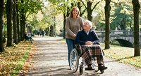Young Caucasian female caregiver pushing elderly Caucasian man in wheelchair along tree-lined park path during autumn season for healthcare assistance concept.