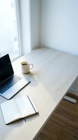 Minimalist workspace with laptop, coffee mug, and open notebook on light wood desk near bright window, ideal for remote work and productivity concepts.