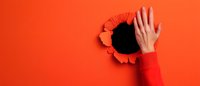 Hand in red sleeve breaking through torn orange paper background creating dramatic hole effect, minimal creative concept for breakthrough and disruption.