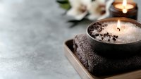 Glowing spa candles with soft flame on dark towel, white orchid flowers in background on grey stone surface. Wellness and relaxation concept.