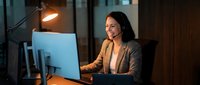 Professional Asian businesswoman working late at office desk with computer and warm desk lamp lighting creating productive evening workspace atmosphere.