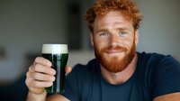 Smiling redhead man with freckles and ginger beard holding glass of green beer in navy t-shirt against blurred background. St Patrick's Day celebration concept.