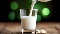 Fresh milk pouring from bottle into glass on rustic wooden table with green bokeh background, dairy drink splash in natural lighting.