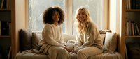 Two diverse women in cozy pajamas having intimate conversation on bed by window. Morning light creates warm atmosphere for friendship bonding moment.