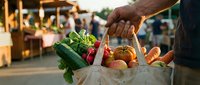 Person holding reusable bag filled with fresh vegetables and fruits at outdoor farmers market during daytime shopping.