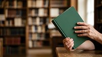 Person holding green hardcover book in library with blurred bookshelves background, suitable for education, reading, literature, and knowledge concepts.