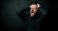 Stressed middle-aged Caucasian man with beard screaming in frustration holding head with hands against dark background expressing mental breakdown.